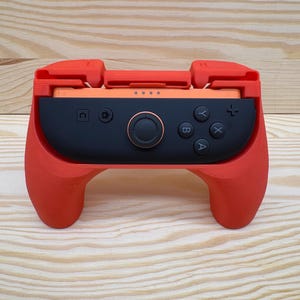 Può includere: Un'impugnatura per controller di videogiochi rossa e nera. Il controller ha un centro nero con pulsanti e un joystick, e maniglie e pezzi superiori rossi. Lo sfondo è una venatura del legno chiara.