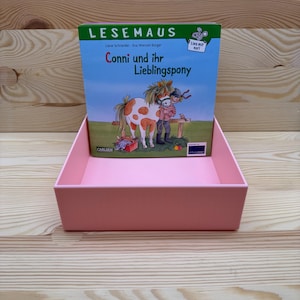 May include: A pink display box holding a children's book titled "Conni und ihr Lieblingspony." The book cover features an illustration of a girl and a pony. The text "LESEMAUS" is at the top. The display box is on a light wood surface.