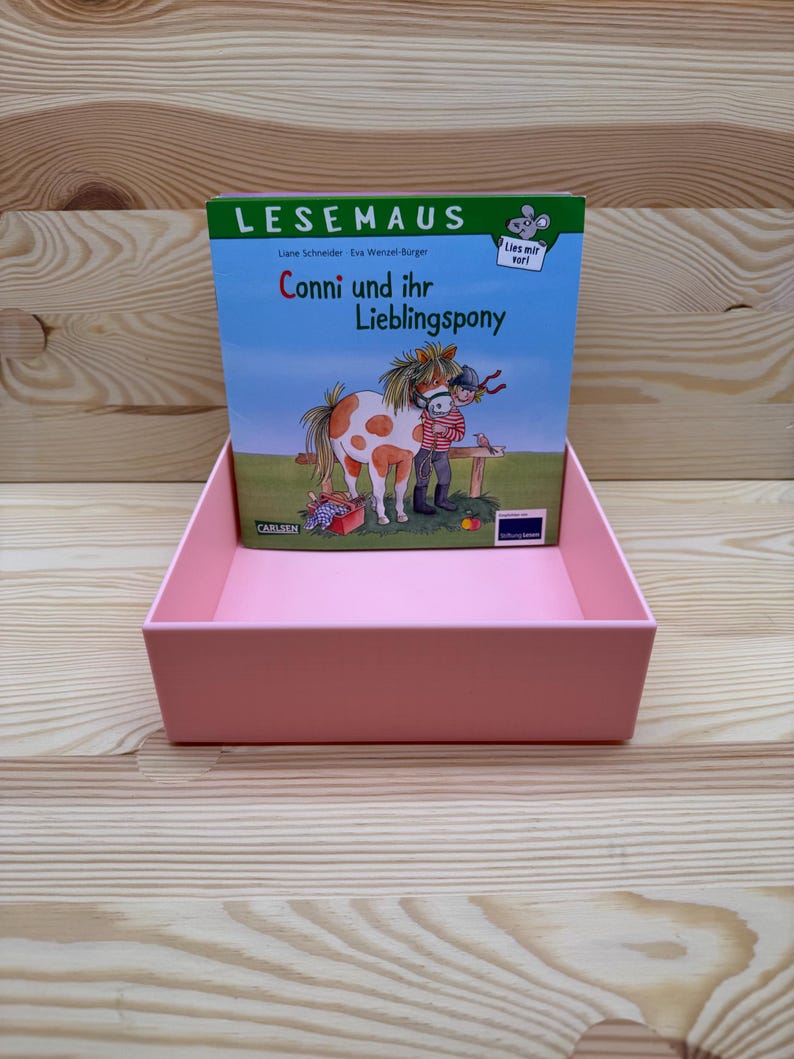 May include: A pink display box holding a children's book titled "Conni und ihr Lieblingspony." The book cover features an illustration of a girl and a pony. The text "LESEMAUS" is at the top. The display box is on a light wood surface.