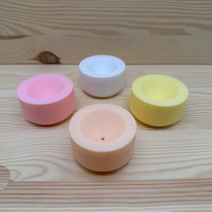 Könnte beinhalten: Vier kleine, runde Behälter in Pastellfarben: Rosa, Weiß, Pfirsich und Gelb. Jeder Behälter hat eine kreisförmige Vertiefung in der Mitte. Sie sind auf einer hellen Holzoberfläche angeordnet. Geeignet für kleine Gegenstände.