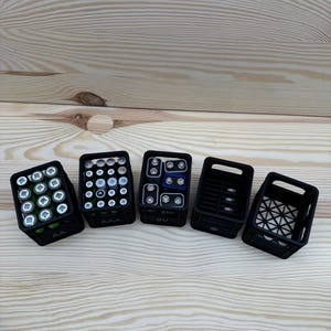 May include: Five black plastic battery organizers filled with various batteries, including AA, AAA, and 9V, are arranged on a light wood surface. The organizers are rectangular and have open tops, showcasing the batteries.