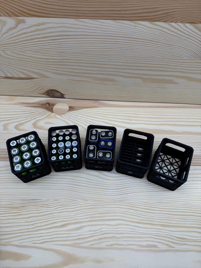 May include: Five black plastic battery organizers filled with various batteries, including AA, AAA, and 9V, are arranged on a light wood surface. The organizers are rectangular and have open tops, showcasing the batteries.