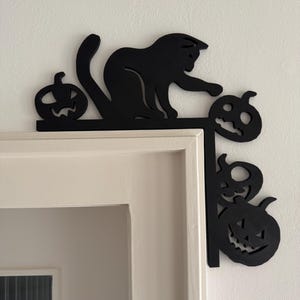 Op de afbeelding: Zwarte Halloween-hoekdecoratie met een kat en uitgesneden pompoenen. De kat zit bovenop een horizontaal stuk en reikt naar een pompoen. Extra pompoenen zijn uitgesneden in het verticale stuk, wat een feestelijke uitstraling geeft.