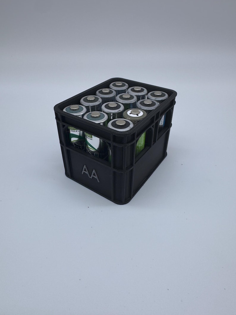 May include: A black plastic crate filled with AA batteries. The batteries are silver and black, and the crate has the letters "AA" printed on the side. The crate is sitting on a white surface.