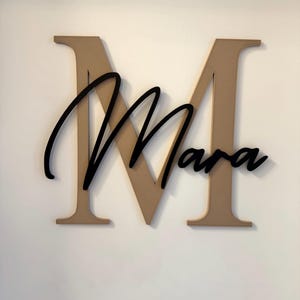 Op de afbeelding: Houten wanddecoratie met een grote, bruine letter "M" met de naam "Mara" in zwart cursief schrift eroverheen. Het bord is op een witte muur gemonteerd, waardoor een contrast ontstaat tussen de donkere en lichte kleuren.
