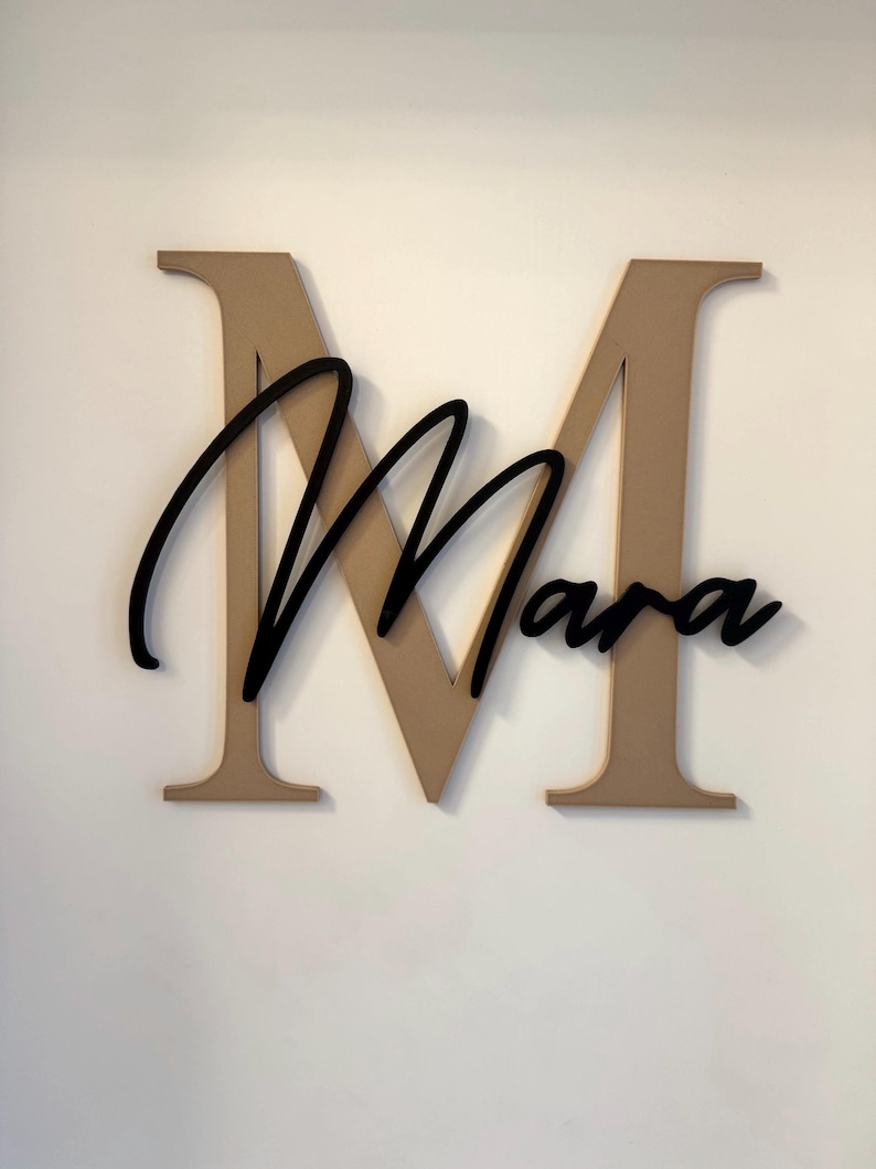 Op de afbeelding: Houten wanddecoratie met een grote, bruine letter "M" met de naam "Mara" in zwart cursief schrift eroverheen. Het bord is op een witte muur gemonteerd, waardoor een contrast ontstaat tussen de donkere en lichte kleuren.