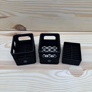 May include: Three black plastic crates of varying sizes are arranged on a light wood surface. The crates have rectangular openings and a grid-like pattern on the bottom. The smallest crate is a solid block.