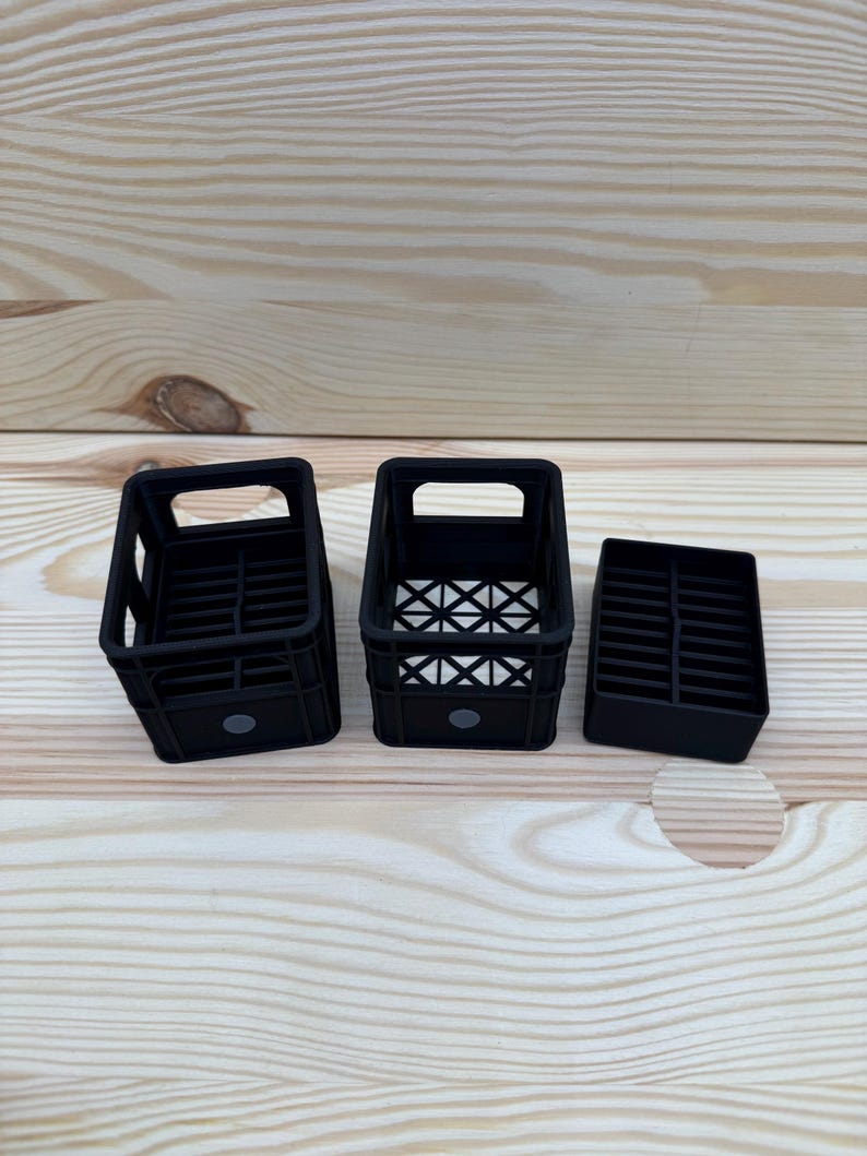 May include: Three black plastic crates of varying sizes are arranged on a light wood surface. The crates have rectangular openings and a grid-like pattern on the bottom. The smallest crate is a solid block.