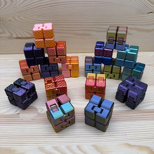 Può includere: Una collezione di colorati cubi fidget in varie tonalità di arancione, viola, blu, verde e bronzo. I cubi sono disposti su una superficie di legno chiaro, mostrando il loro design geometrico e la finitura metallica. Questi giocattoli sono progettati per alleviare lo stress.