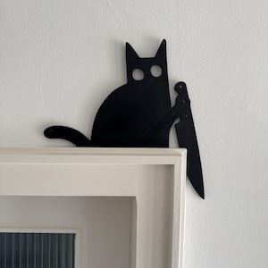 Puede incluir: Silueta negra de un gato sosteniendo un cuchillo, colocada sobre un marco de puerta blanco. El gato tiene ojos grandes y redondos y orejas puntiagudas. El cuchillo también es negro, creando un diseño humorístico y ligeramente amenazante.