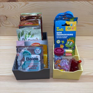 Könnte beinhalten: Zwei Kunststoff-Organizer auf einer Holzoberfläche. Ein grauer Organizer enthält Päckchen und eine Schachtel Balea Hydrogel Augen Pads. Der andere gelbe Organizer enthält ein Tinti-Badeprodukt, einen rosa Behälter und kleine Päckchen.
