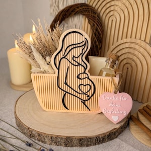 Könnte beinhalten: Ein Holzkorb mit einer Silhouette einer schwangeren Frau, gefüllt mit Kerzen und getrockneten Pflanzen. Eine kleine Flasche und ein rosa herzförmiges Etikett mit Text sind ebenfalls vorhanden. Die Anordnung befindet sich auf einer Holzscheibe.