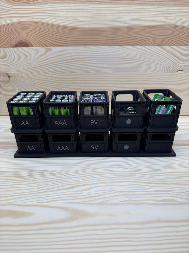 May include: A black battery organizer with multiple compartments, each labeled for different battery types: AA, AAA, and 9V. The organizer is filled with various batteries, providing a neat storage solution.