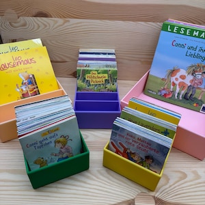 Op de afbeelding: Een verzameling kinderboeken in kleurrijke dozen. De boeken hebben Duitse titels, waaronder "Leo", "Conni" en "Das Wildschwein-Picknick". De dozen zijn in verschillende kleuren: perzik, paars, groen, geel en roze.