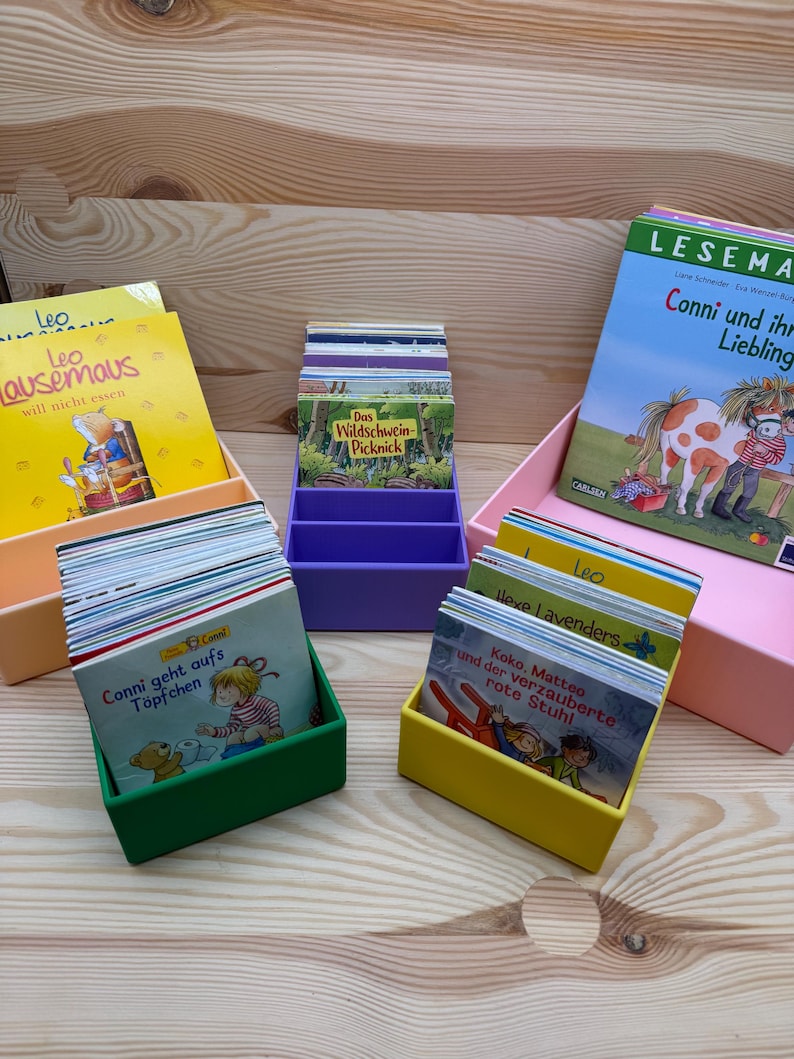 May include: A collection of children's books in colorful boxes. The books have titles in German, including "Leo", "Conni", and "Das Wildschwein-Picknick". The boxes are in various colors: peach, purple, green, yellow, and pink.