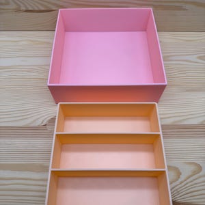 May include: Two-piece pink storage box set. The larger box is a solid pink, while the smaller box has three compartments in a lighter shade of pink. The boxes are made of a smooth material and are set against a light wood grain background.