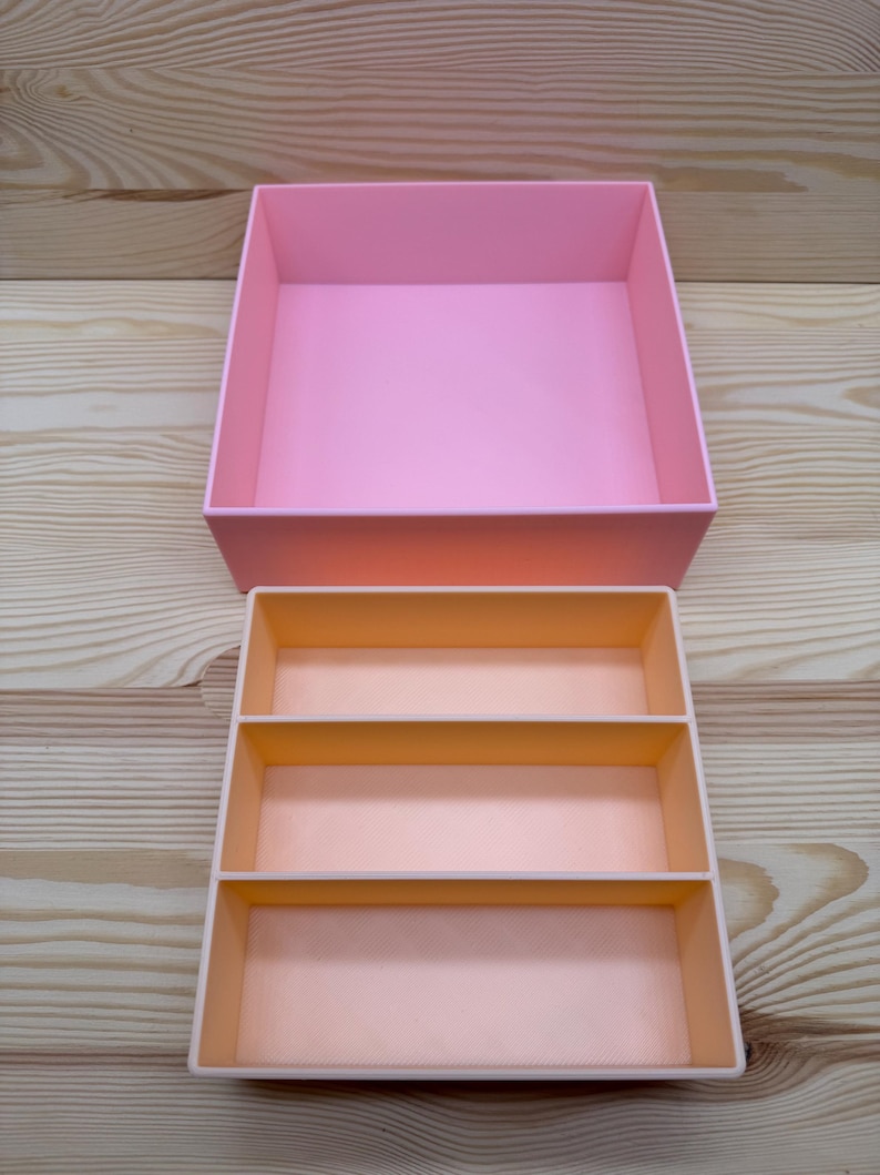 May include: Two-piece pink storage box set. The larger box is a solid pink, while the smaller box has three compartments in a lighter shade of pink. The boxes are made of a smooth material and are set against a light wood grain background.