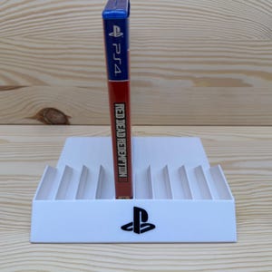 Può includere: Un supporto bianco per giochi PS4 con un logo PlayStation nero contiene una custodia del gioco Red Dead Redemption II. La custodia del gioco è rossa e presenta il testo "Red Dead Redemption II" e "PS4". Il supporto ha più fessure.