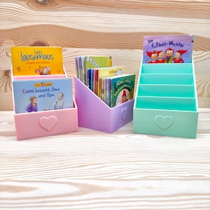 Può includere: Tre organizzatori di libri colorati in rosa, viola e verde menta, ciascuno con un ritaglio a forma di cuore. Contengono libri per bambini con titoli come "Leo Lausemaus" e "Conni besucht Oma und Opa". Lo sfondo è in legno chiaro.