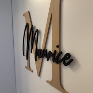 Op de afbeelding: Een aan de muur gemonteerd bord met een grote, goudkleurige letter "M" met de naam "Maurice" in zwart script eroverheen. Het bord is gemaakt van hout en is aan een witte muur bevestigd.