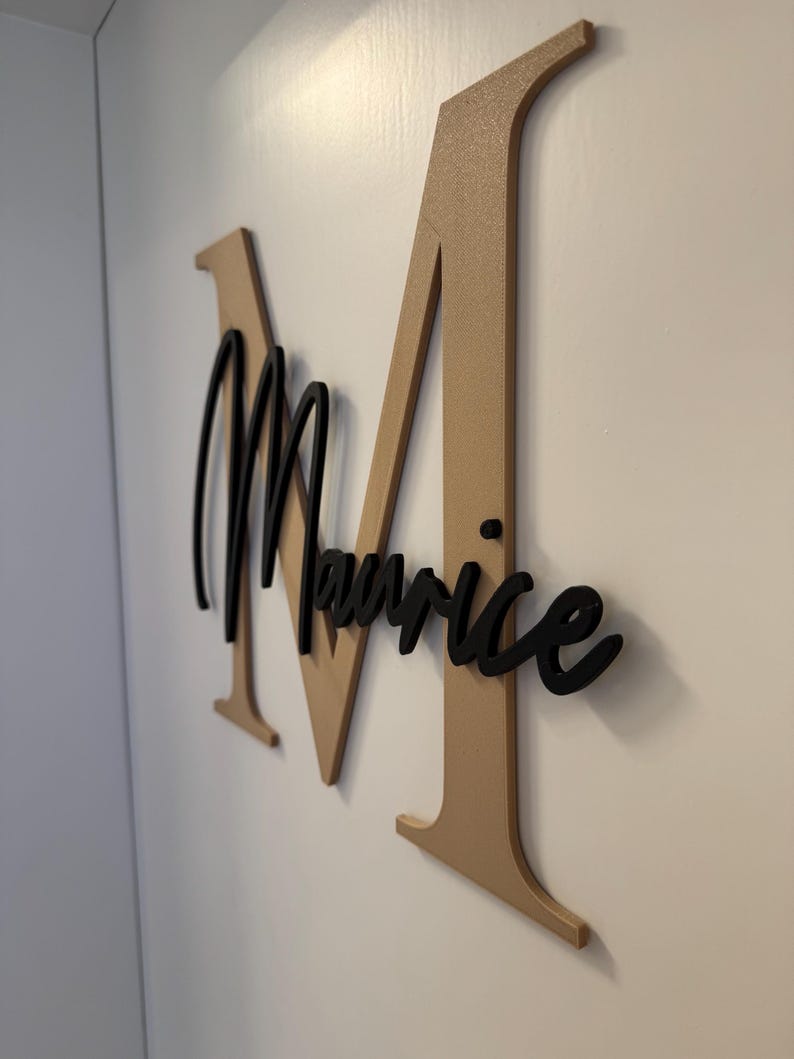 Op de afbeelding: Een aan de muur gemonteerd bord met een grote, goudkleurige letter "M" met de naam "Maurice" in zwart script eroverheen. Het bord is gemaakt van hout en is aan een witte muur bevestigd.