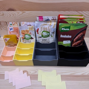 Könnte beinhalten: Eine Auswahl an Lebensmittelpackungen und Behältern, darunter Vanillezucker, Bourbon-Vanille und Vanillepudding. Die Packungen sind in pastellfarbenen Organisatoren angeordnet, mit zusätzlichen gelben und rosa Trennwänden.