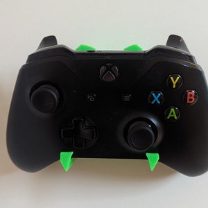 May include: A black Xbox controller with green plastic wall mounts. The controller has the standard Xbox buttons: A, B, X, Y, and a directional pad.