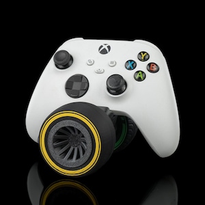 Pode incluir: Um controlador Xbox branco com um acessório de aderência em forma de pneu preto e cinza. O pneu tem uma faixa amarela ao redor da borda.