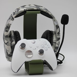 Pode incluir: Um controlador de jogo branco e um fone de ouvido camuflado com microfone são exibidos em um suporte verde.