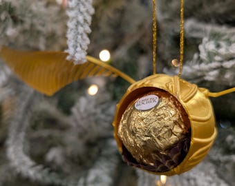 May include: A golden Snitch ornament with a Ferrero Rocher candy inside, hanging from a Christmas tree. The ornament has wings and is suspended by a gold string. The tree is decorated with lights and has a frosted appearance.