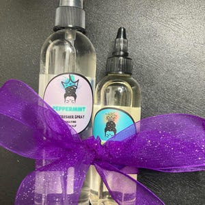 Peut inclure: Deux flacons transparents avec des vaporisateurs noirs et des étiquettes. Le plus grand flacon a une étiquette violette et blanche avec le texte "Peppermint Fresher Spray" et une image de dessin animé d'une femme avec une couronne. Le plus petit flacon a une étiquette blanche et bleue avec une image de dessin animé d'une femme avec un ananas sur la tête. Les deux flacons sont liés ensemble par un ruban violet.