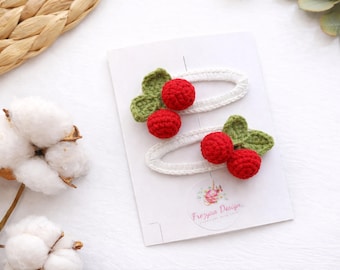 Puede incluir: Dos pinzas para el pelo de ganchillo blancas, cada una adornada con un par de cerezas rojas de ganchillo y hojas verdes. Las pinzas se muestran en una tarjeta blanca con el logotipo "Frezyna Design".