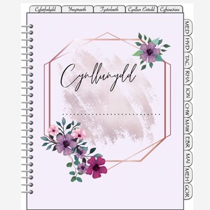 May include: A purple planner with a floral design and a gold octagon frame. The planner has a spiral binding and tabs for each month of the year. The text "Cynllunydd" is written in black on the front cover.