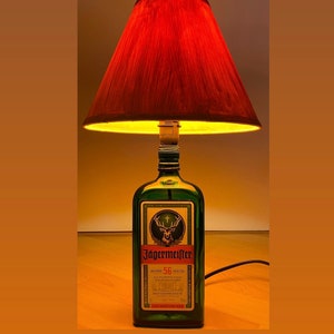 Puede incluir: Una lámpara de botella de vidrio verde con una pantalla roja. La botella está etiquetada con "Jägermeister 56 Kräuter", con un logotipo de ciervo y cruz. La pantalla está hecha de tela y tiene un patrón de veta de madera.