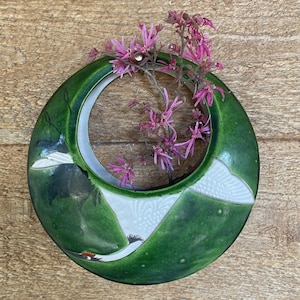 May include: A green ceramic vase with a circular design, featuring a white and gray mountain scene with a crane. The vase holds pink flowering branches. The vase is approximately 8 inches in diameter.