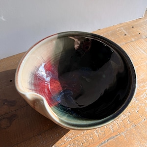 May include: A ceramic bowl with a glossy finish featuring a deep black interior that transitions into a vibrant red and dark green gradient on the outer rim. The bowl has a distinct pouring spout integrated into its circular, curved edge.