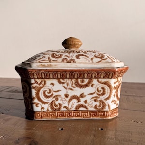 May include: A rectangular ceramic lidded container featuring a white base with intricate brown floral scrollwork patterns. The lid has a central knob, and the base is decorated with a brown Greek key border design along the bottom edge.