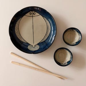 May include: A ceramic dinnerware set consisting of one large round plate and two small bowls, all featuring a cream-colored center with a dark blue fish illustration. Two light-colored wooden chopsticks are positioned diagonally below the plate.