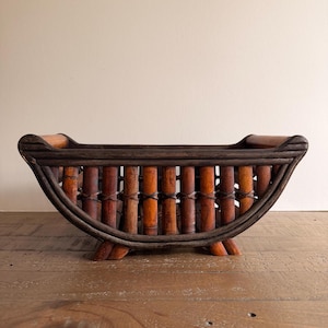 May include: A curved, boat-shaped basket constructed from dark brown bamboo slats. The structure features vertical bamboo rods secured with thin, dark woven bindings, creating an open-slatted design with a sturdy base and two small feet.