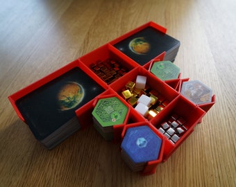 May include: Red plastic organiser with compartments for storing game pieces. The organiser has two stacks of cards with images of planets, a stack of green hexagonal tiles, a stack of blue hexagonal tiles, a pile of gold and silver cubes, and a pile of small silver squares.