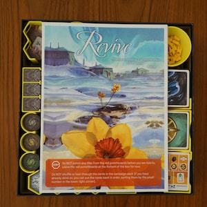 May include: A board game box with a blue and yellow illustration of a daffodil on the cover. The game is called "Revive: The Reclamation of a Lost World".