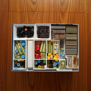 May include: A white organizer with multiple compartments filled with various game pieces, including wooden sticks, small colored cubes, and cards. The organizer is ideal for storing and organizing game components.