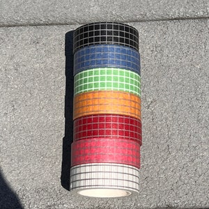 May include: A stack of seven rolls of decorative washi tape in various colours. The colours include black, blue, green, orange, red, pink, and white. Each roll has a grid pattern.
