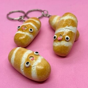 May include: Three small, white and brown, baguette-shaped keychains with googly eyes and pink noses. The keychains are on a pink background.