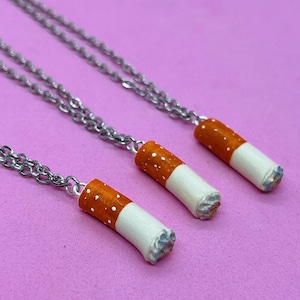 May include: Three silver chain necklaces with a small, white and orange cigarette charm on each. The necklaces are on a pink background.