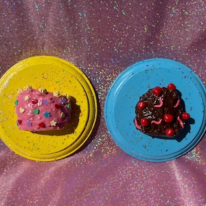 May include: Two miniature cakes on round plates. One cake is pink with colorful star and heart-shaped sprinkles on a yellow plate. The other cake is dark brown with red cherries and pink icing swirls on a light blue plate.