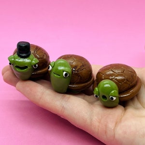 May include: Three small, hand-crafted, brown and green turtle figurines. The turtle on the left is wearing a black top hat.