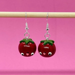 May include: A pair of red tomato earrings with green stems and white eyes. The earrings are made of polymer clay and have a cute, cartoonish look.