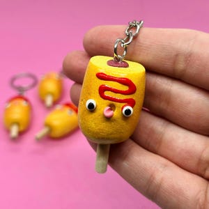May include: A yellow and red corndog keychain with googly eyes and a pink mouth. The corndog is on a wooden stick and has a silver chain attached to it.