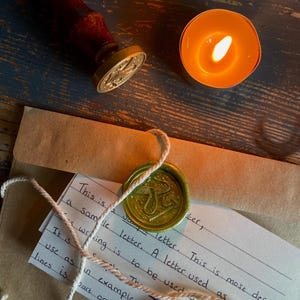 Puede incluir: Una vela encendida junto a un sobre marrón con un sello de cera verde y un sello de madera. El sello tiene un diseño de hongo. Se ve una nota manuscrita con el texto: "This is a sample letter."